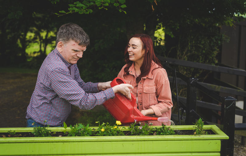 A man is watering flowers while a woman is smiling beside him